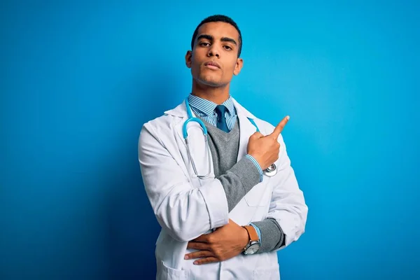 一位英俊的非洲裔美国医生 身穿外套 带着听诊器 头戴蓝色背景 手指头指向广告的侧面 严肃而沉着 — 图库照片