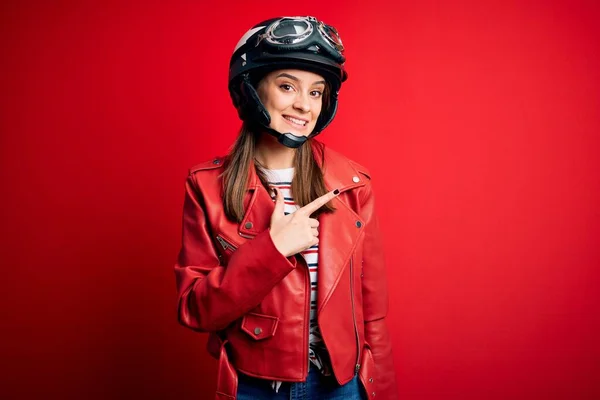 若い美しいブルネットのオートバイのヘルメットと顔の笑顔で陽気に赤いジャケットを身に着けている自転車愛好家の女性が顔に幸せと自然な表情で側に手と指で指して — ストック写真