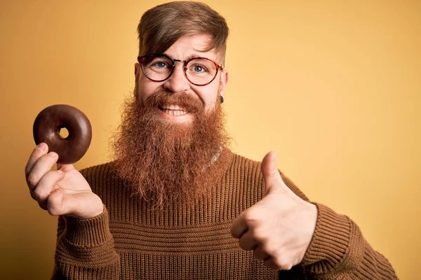 Redhead Irish man with beard eating chocolate donut over yellow background happy with big smile doing ok sign, thumb up with fingers, excellent sign