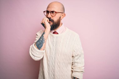 Sakallı, dövmeli, pembe arka planda gözlüklü ve kazaklı yakışıklı, gergin ve ağzı tırnak yiyen elleri olan bir adam. Endişe sorunu.