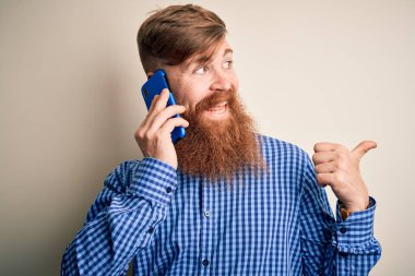 Kızıl saçlı, sakallı İrlandalı bir adam akıllı telefondan konuşuyor izole bir arka plan üzerinden işaret ediyor ve gülümseyen bir yüzle baş parmağıyla gösteriyor.