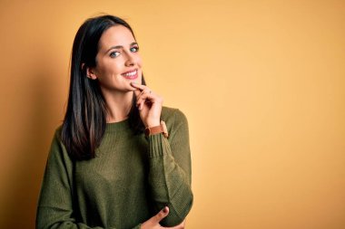 Sarı arka planda yeşil süveter giyen, mavi gözlü genç esmer kadın çapraz kollarla gülümseyen ve elleri çenesinde yükselen kameraya kendinden emin bir şekilde bakıyor. Olumlu düşünüyorum..
