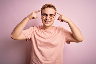 Genç, yakışıklı, günlük tişört giyen, pembe arka planda dikilen, iki eliyle başını gösteren, harika bir fikir ya da düşünce, iyi bir hafıza.