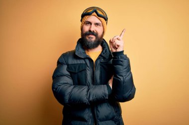 Sakallı, kel, yakışıklı kayakçı kar bezi giymiş, yüzünde kocaman bir gülümsemeyle kayak gözlüğü takmış, kameraya el ve parmak şeklinde bakıyor..