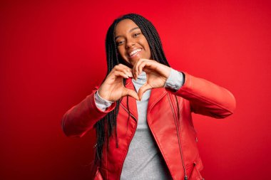Genç Afro-Amerikalı kadın, kırmızı izole edilmiş arka planda havalı bir deri ceket giyiyor. Elleriyle kalp sembolü yapan bir aşkla gülümsüyor. Romantik konsept.