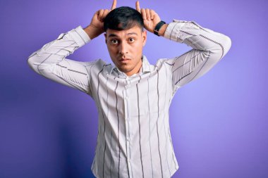 Zarif iş gömleği giyen genç yakışıklı İspanyol adam mor arka planda duruyor ve boynuz gibi parmağıyla komik jestler yapıyor.