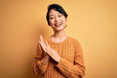 Genç, güzel Asyalı kız gündelik süveter ve Diadem giyip sarı arka planda duruyor alkışlıyor ve mutlu, neşeli ve gururlu elleri alkışlıyor.