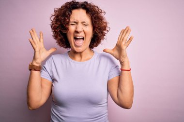 Orta yaşlı güzel kıvırcık saçlı kadın pembe arka plan üzerine gündelik tişört giyip başarının çılgınlığını kollarını kaldırarak ve gözlerini kapayarak heyecanla bağırarak kutluyor. Kazanan konsept