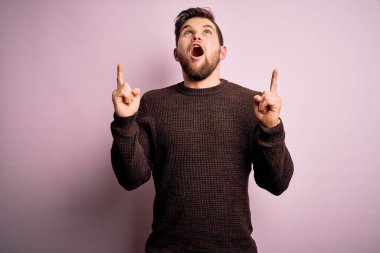 Sarışın, sakallı ve mavi gözlü genç adam pembe arka planda sıradan bir süveter giyiyor şaşkın şaşkın bakıyor, parmaklarıyla işaret ediyor ve kollarını kaldırıyordu..
