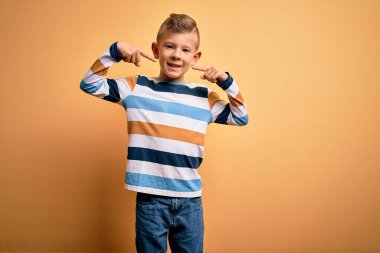 Mavi gözlü, sarı arka planda çizgili tişört giyen, neşeli gülümseyen, parmakları, dişleri ve ağzıyla işaret eden küçük beyaz bir çocuk. Diş sağlığı konsepti.