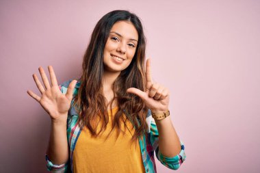 Genç, güzel, esmer, sıradan bir gömlek giyen, pembe arka planda duran, yedi numarayı gösteren, kendinden emin ve mutlu bir şekilde gülümseyen bir kadın..