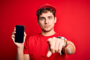 Kıvırcık saçlı, şapkalı, kırmızı arka planda akıllı telefon tutan, kamerayı işaret eden genç, sarışın teslimatçı. Ve sen, el işareti, ön taraftan pozitif ve kendinden emin bir hareket.