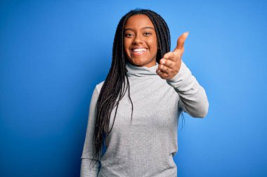 Genç Afro-Amerikalı kadın mavi izole edilmiş arka plan üzerine balıkçı yaka kazak giyip dostça bir selamlama ve hoş geldin hediyesi olarak el sıkışmayı teklif ediyor. Başarılı bir iş..