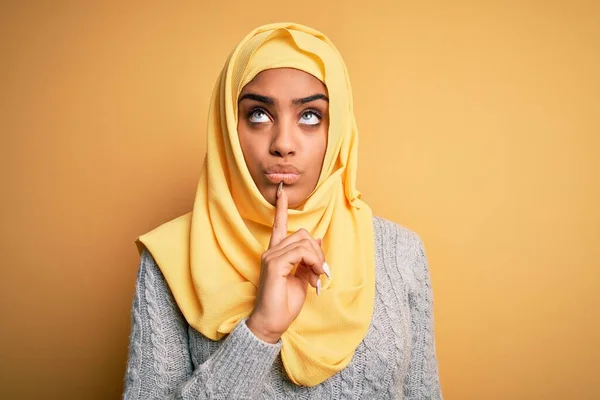 Young beautiful african american girl wearing muslim hijab over isolated yellow background Thinking concentrated about doubt with finger on chin and looking up wondering