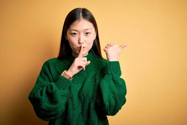 年轻美丽的亚洲女人穿着绿色冬季毛衣 披着黄色孤立的背景 要求保持安静 手指放在嘴唇上 手拉手指向侧面 沉默和秘密概念 — 图库照片