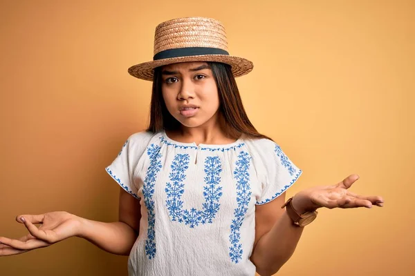 年轻美丽的亚洲女孩穿着休闲装 戴着一顶帽子 站在黄色的背景上 毫无头绪 与张开的双臂混淆不清 没有概念 — 图库照片