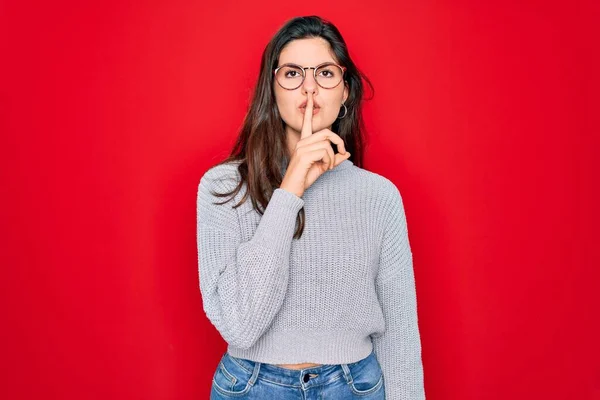 若い美しいブルネットの女性は 唇に指で静かになるように求めている赤い背景にカジュアルなセーターを着ています 沈黙と秘密の概念 — ストック写真
