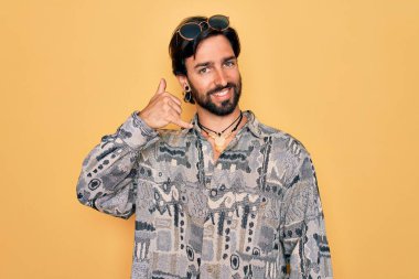 Yakışıklı İspanyol bohem bir adam hippi tarzı giyinmiş ve güneş gözlüklü gülümsüyor telefonla el ve parmaklarıyla telefonla konuşuyor gibi yapıyor. İletişim kavramları.