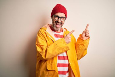 Orta yaşlı, ağarmış, gözlüklü, yağmurluk takan, izole edilmiş beyaz arka planda duran, gülümseyen ve kameraya bakan, iki eli ve parmakları yanda olan bir adam..