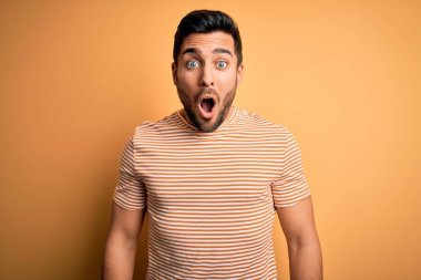 Sakallı, sarı arka planda sıradan çizgili t-shirt giyen genç yakışıklı bir adam korku, korku ve heyecan dolu bir yüz ifadesiyle korkmuş ve şok olmuştu..