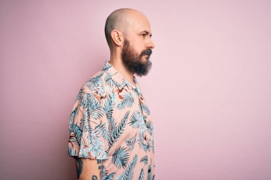 Sakallı ve dövmeli yakışıklı, pembe arka planda sıradan bir çiçek gömleği giyen, rahat bir görünüm takınan, doğal yüzlü ve kendine güvenen bir gülümseme takınan kel bir adam..