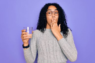Genç Afrikalı Amerikalı kadın, izole edilmiş mor arka planda bir bardak su içiyor. Ağzı, hata yüzünden utançla kaplı, korkunun ifadesi, sessizlikten korkmak, gizli bir konsept.