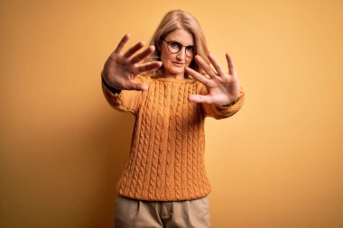 Orta yaşlı güzel sarışın kadın sarı arka planda günlük süveter ve gözlük takıyor. El ve parmakları kullanarak çerçeve yapıyor.