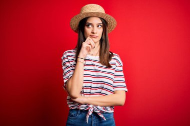 Genç, güzel, esmer, çizgili tişört ve kırmızı arka planda yaz şapkası giyen, eli çenesinde soru soran, düşünceli bir kadın. Düşünceli bir yüzle gülümsüyor. Şüphe kavramı.