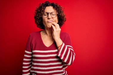 Orta yaşlı güzel kıvırcık saçlı kadın diş ağrısı ya da diş hastalığından dolayı kırmızı arka planda sıradan çizgili süveter giyip dudaklarına acı dolu bir ifadeyle dokunuyor. Dişçi