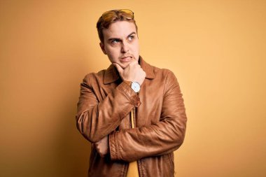 Sarı arka planda sıradan deri ceket giyen genç ve yakışıklı bir adam bir soru hakkında endişeli ve gergin bir şekilde düşünüyor.