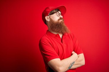 Gözlüklü ve kırmızı şapkalı genç teslimatçı kollarını kavuşturmuş ve kendinden emin bir şekilde kollarını yana bakıyor.