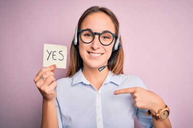 Güzel çağrı merkezi ajanı kadın kulaklık takarak çalışıyor. Elinde evet mesajı ve kendini işaret eden sürpriz bir yüz ifadesi var.