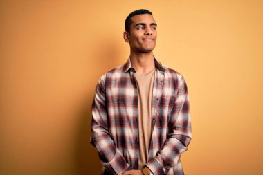 Genç, yakışıklı, Afro-Amerikan bir adam, günlük gömleğini giymiş sarı arka planda dikiliyor, gülümsüyor ve başka tarafa bakıyor..