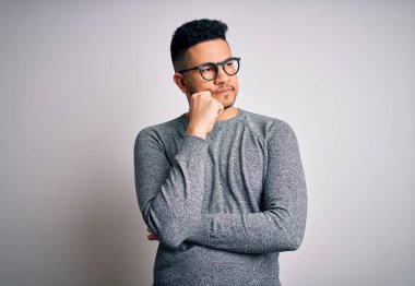 İzole edilmiş beyaz arka plan üzerinde gözlük takan genç yakışıklı bir adam. Elleri çenesinde soru sorarken, düşünceli bir ifade takınıyor. Düşünceli bir yüzle gülümsüyor. Şüphe kavramı.