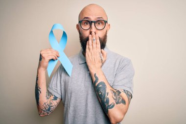 Sakallı ve dövmeli yakışıklı kel bir adam mavi kanser kurdelesini izole arka planda tutuyor. Ağzı kapalı. Yanlışlıkla utançla dolu.