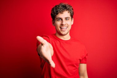 Sarışın, kıvırcık saçlı, kırmızı arka planda sıradan tişörtler giyen, neşeli gülümsemeli, yardım eli uzatan ve kabul gören genç bir adam..