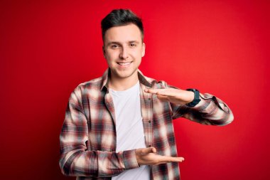 Kırmızı izole edilmiş arka plan jestlerinin üzerine gündelik modern tişörtler giyen genç, yakışıklı beyaz bir adam. Elleri büyük ve büyük boy işareti, ölçü sembolü gösteriyor. Kameraya bakıp gülümsüyor. Ölçüm konsepti.