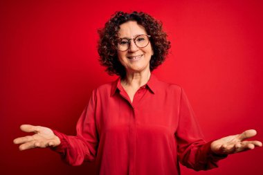 Orta yaşlı güzel kıvırcık saçlı kadın kırmızı arka planda günlük gömlek ve gözlük takıyor neşeli gülümseyerek yardım eli uzatıyor ve kabul ediyor..