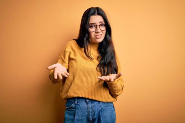 Genç esmer kadın gözlük takıyor ve sarı izole arka planda sıradan bir süveter giyiyor. Hiçbir fikri yok ve kolları açık, hiçbir fikri yok..