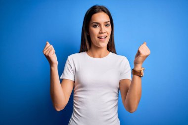 Genç, güzel, esmer, beyaz tişörtlü bir kadın mavi arka planda dikilmiş, kollarını kaldırarak ve açık gözlerle başarıyı kutluyor. Kazanan konsept.