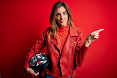 Güzel esmer motosikletli kadın kırmızı ceketli motosiklet kaskı tutan mutlu bir şekilde elini ve parmağını kenarı işaret ediyor.