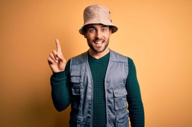 Tatilde sakallı yakışıklı turist sarı arka planda kaşif şapkası takıyor ve başarılı bir fikri işaret ediyor. Heyecanlı ve mutlu. Bir numara..