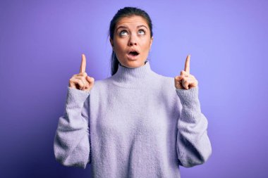 Mavi gözlü genç bir kadın pembe arka planda balıkçı yaka kazak giyiyor şaşkın şaşkın yukarı bakıyor ve parmaklarıyla işaret ediyor ve kollarını kaldırıyor..