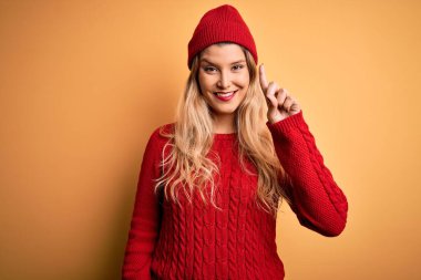 Beyaz arka planda günlük süveter ve yün şapka giyen genç, güzel sarışın kadın bir numaralı parmağını gösterirken kendinden emin ve mutlu bir şekilde gülümsüyor..