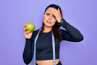 Güzel esmer sporcu kadın, bant mezurası kullanıyor mor arka planda elma meyvesi yiyor gergin ve hayal kırıklığına uğramış, şaşırmış ve sinirli bir yüz ifadesi var.