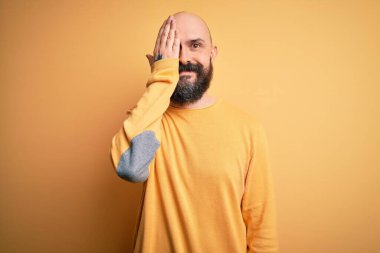 Sarışın, kel, sakallı bir adam sarı arka planda dikiliyor. Bir gözünü eliyle kapatıyor. Yüzünde kendine güvenen bir gülümseme ve sürpriz bir duygu var..