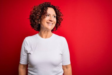 Orta yaşlı güzel kıvırcık saçlı kadın izole edilmiş kırmızı arka plan üzerine günlük tişört giyip yüzünde gülümseme, doğal bir ifade ile yan yana bakıyor. Kendinden emin gülen.