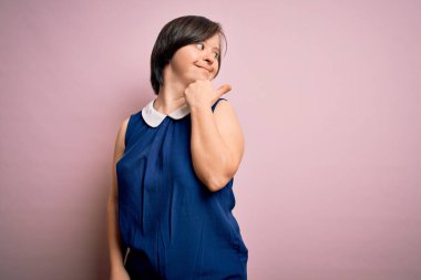 Genç Down sendromlu kadın pembe arka planda zarif bir gömlek giyiyor. Gülümsüyor, gülümsüyor ve başparmağıyla yan tarafa bakıyor..