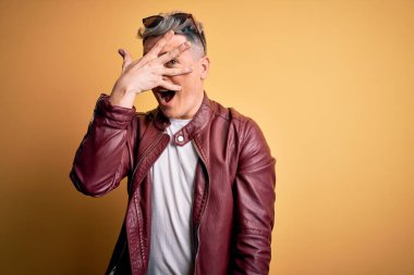 Sarı arka plan üzerinde moda deri ceket ve güneş gözlüğü takan genç, yakışıklı, modern bir adam. Şok olmuş bir şekilde yüzünü ve gözlerini kapıyor, utanmış bir ifadeyle parmaklarından bakıyor..