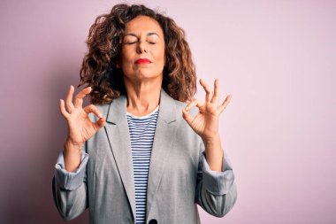 Soyutlanmış pembe arka planda zarif bir ceket giyen orta yaşlı güzel iş kadını rahatla ve parmaklarıyla meditasyon hareketi yaparken gözleri kapalı gülümse. Yoga konsepti.